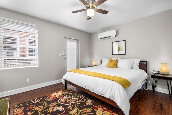 Un dormitorio con cama, ventilador de techo, ventana con persianas, una puerta y un equipo de aire acondicionado.