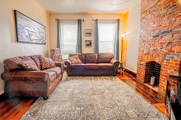 A living room with a brick fireplace, two sofas, a patterned rug, and framed artwork on the wall.