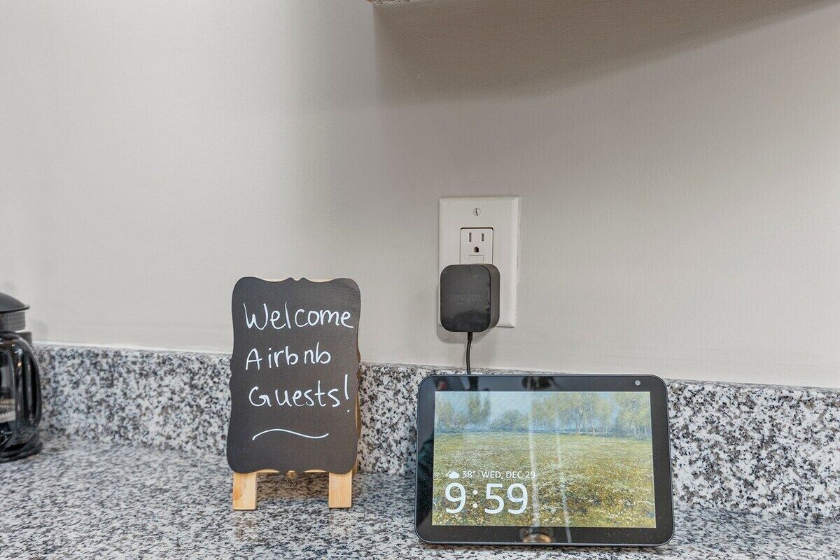 A welcome sign for Airbnb guests, a tablet displaying the date and time, and a power outlet on a wall.