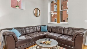 A brown leather sectional sofa with a round coffee table in front, a mirror on the wall, and a window with a view of a brick building. - Classic Soulard 2BD King Master & Private Deck (St. Louis)