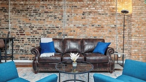 Un salon avec un mur en briques, un canapé en cuir marron agrémenté de coussins bleus, une table basse en verre et des fauteuils bleus.