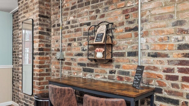 Un mur de briques avec une table en bois et deux chaises. Un panneau noir est accroché au mur, et une petite étagère porte un vase rempli de fleurs.