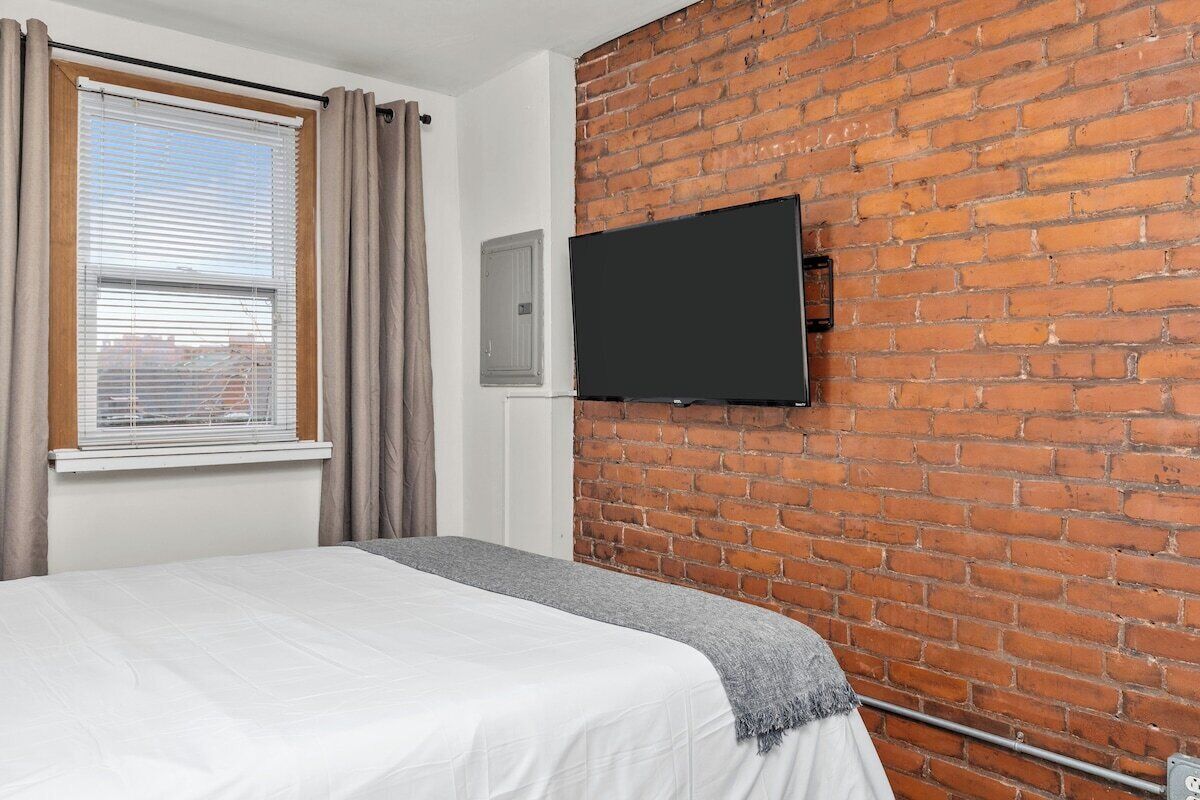 Une chambre d’hôtel avec un mur en briques, un lit recouvert d’une courtepointe blanche, une fenêtre avec des rideaux et un téléviseur à écran plat fixé au mur.
