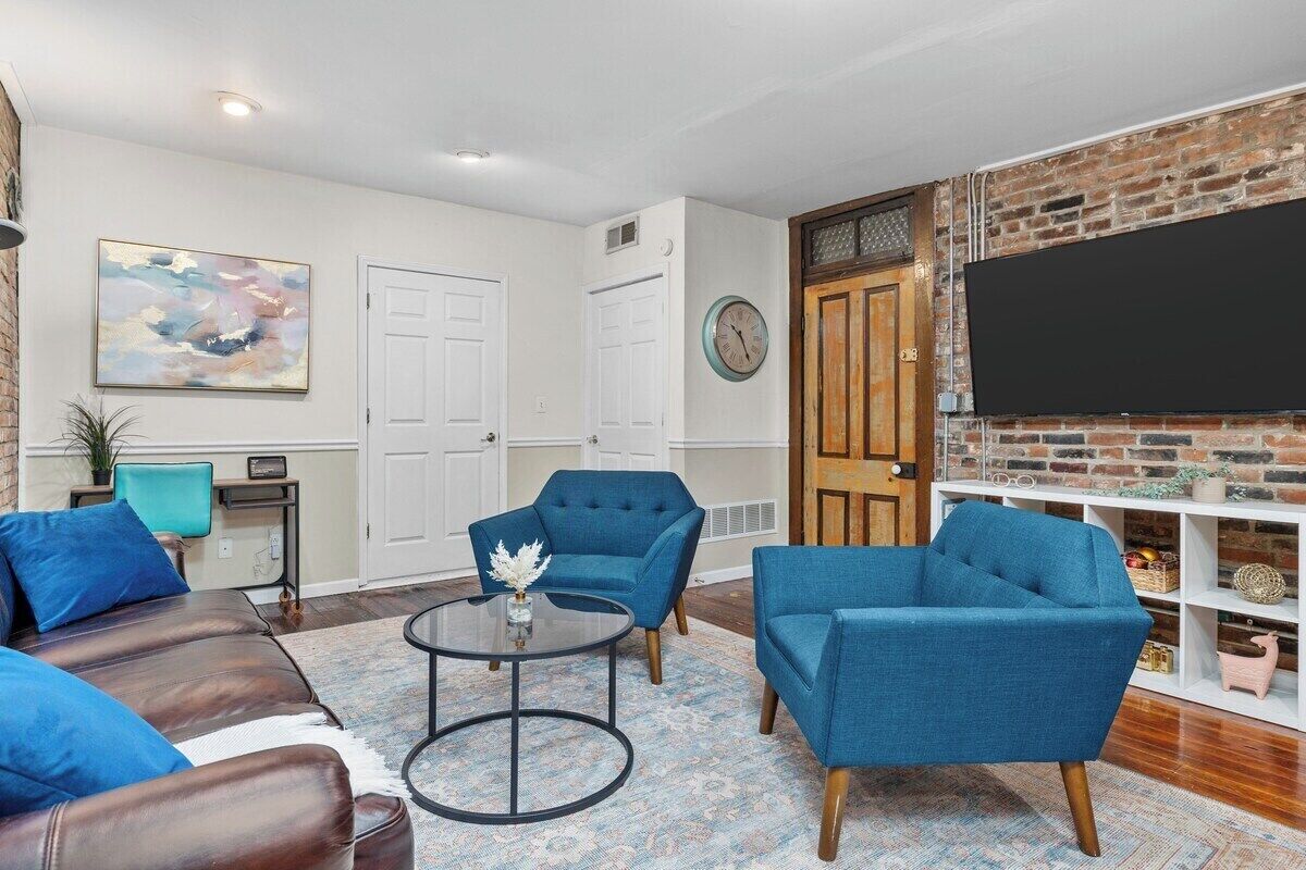 A living room with a brick wall, a flat-screen TV, two blue armchairs, a brown leather sofa, and a round coffee table.