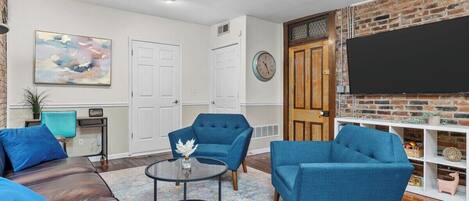 A living room with a brick wall, a flat-screen TV, two blue armchairs, a brown leather sofa, and a round coffee table.
