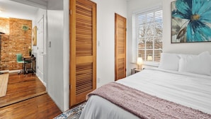 Une chambre avec un grand lit, des portes en bois, une fenêtre avec des stores et un tableau au mur.