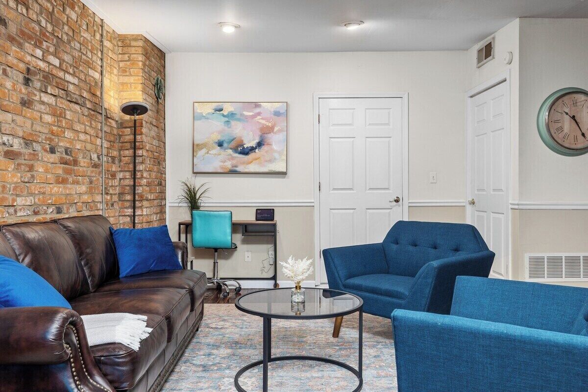 A living room with a brick wall, a blue sofa, a blue armchair, a round coffee table, and a painting on the wall.