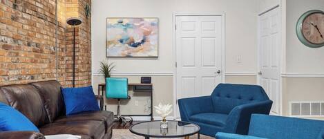 A living room with a brick wall, a blue sofa, a blue armchair, a round coffee table, and a painting on the wall.