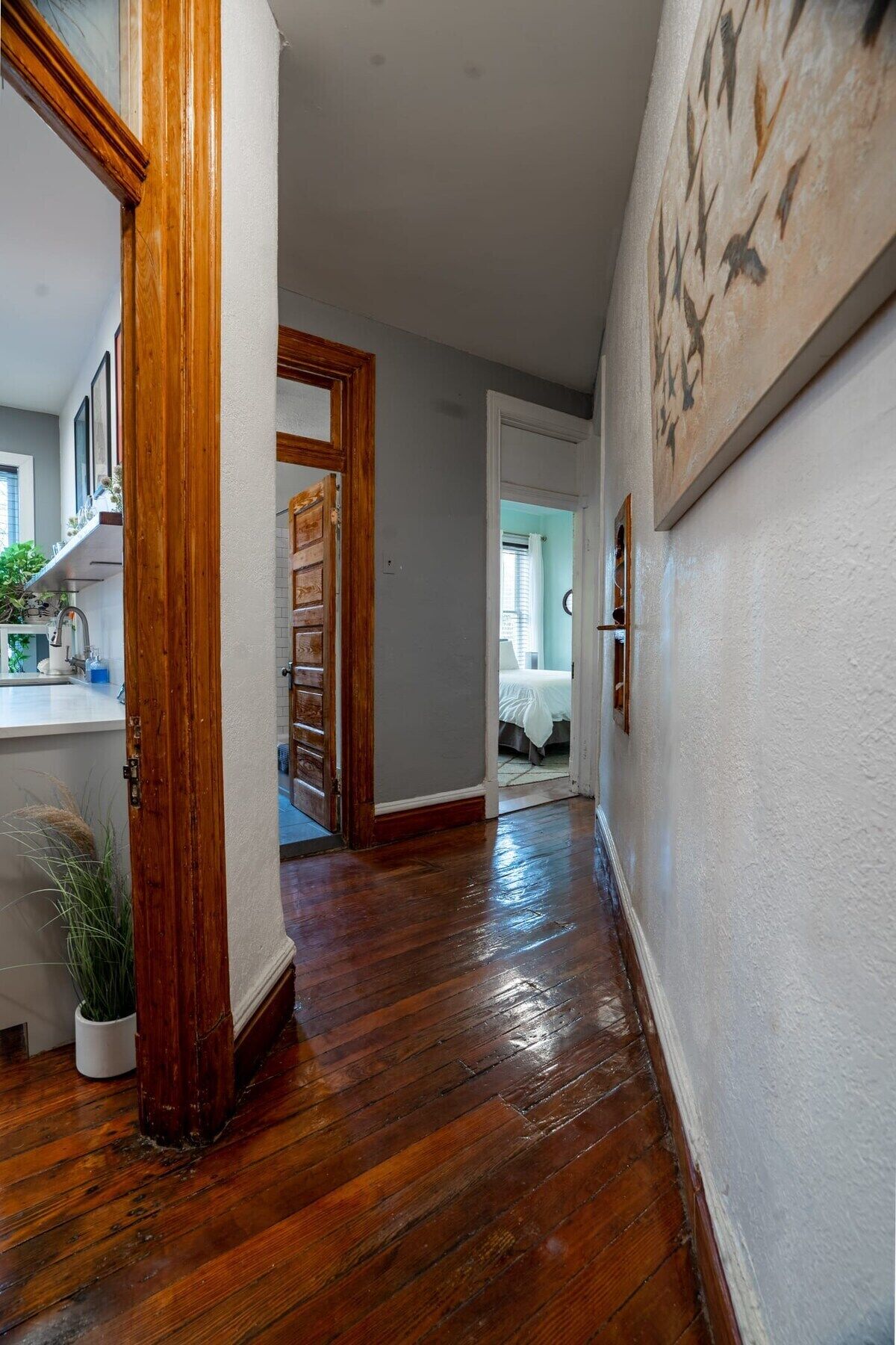 Un couloir au plancher de bois, une porte blanche donnant sur une chambre, et un coin cuisine visible au fond.