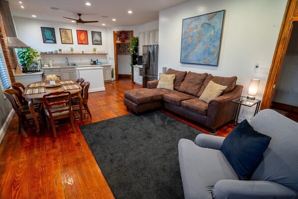 A modern living room with a sectional sofa, a dining table with chairs, a kitchen area, and a large abstract painting on the wall.