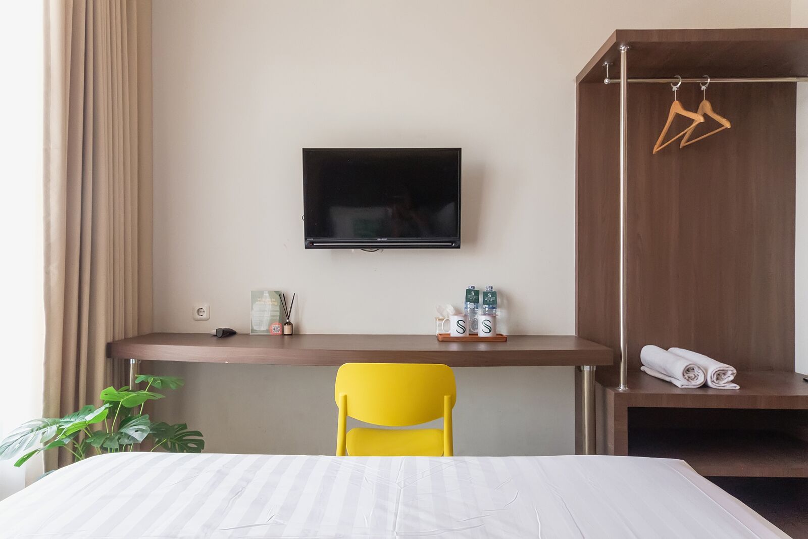 A hotel room with a bed, a desk with a chair, a TV mounted on the wall, a wardrobe with hangers, and a potted plant.