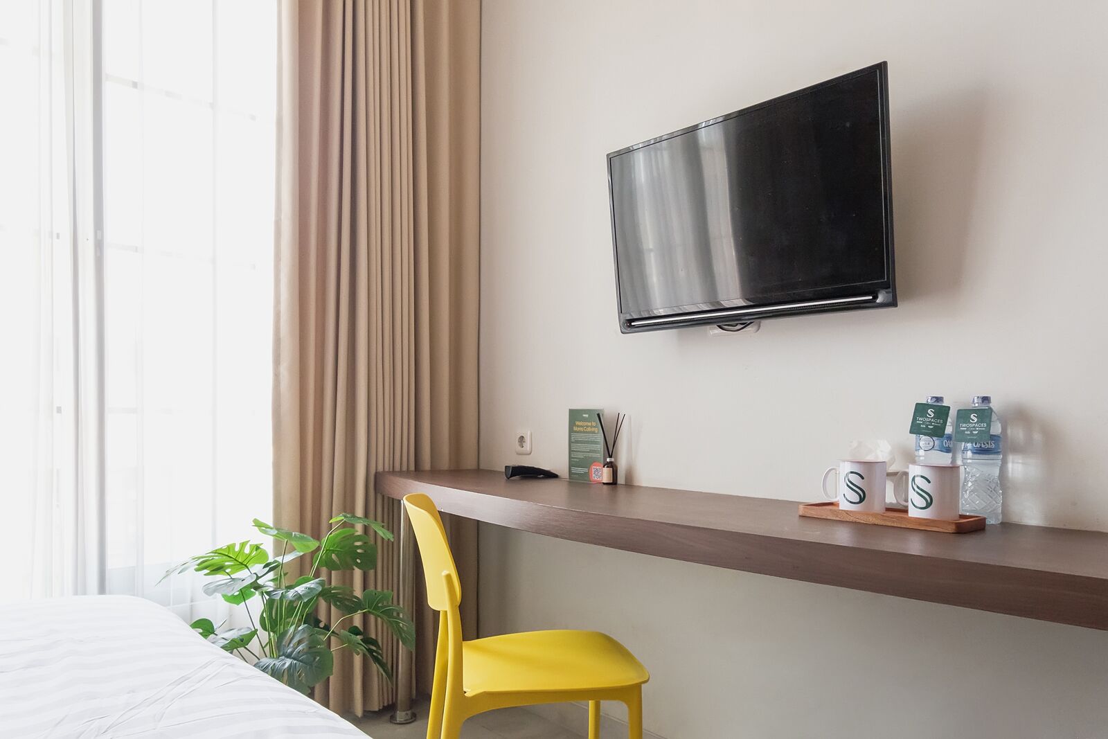 A hotel room with a bed, a desk with a chair, a TV, a plant, and a shelf with water bottles and a sign.