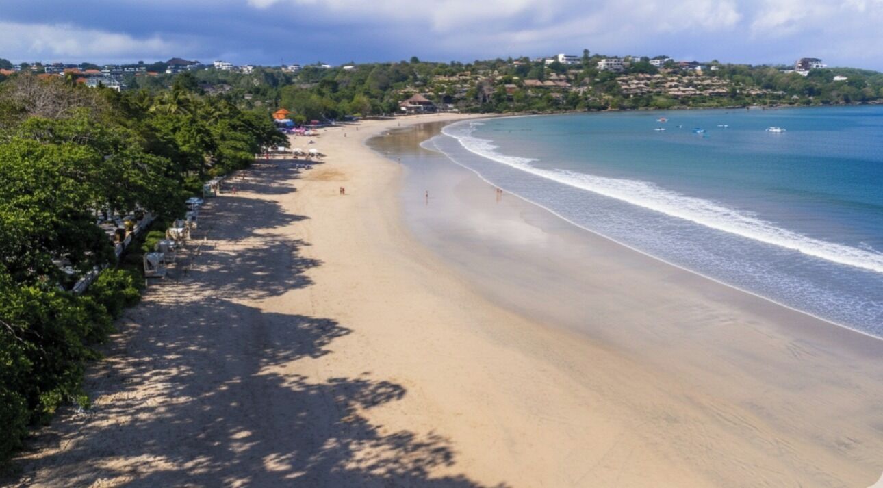 A beach with a clear view of the shoreline, a paved walkway, and a distant view of buildings.