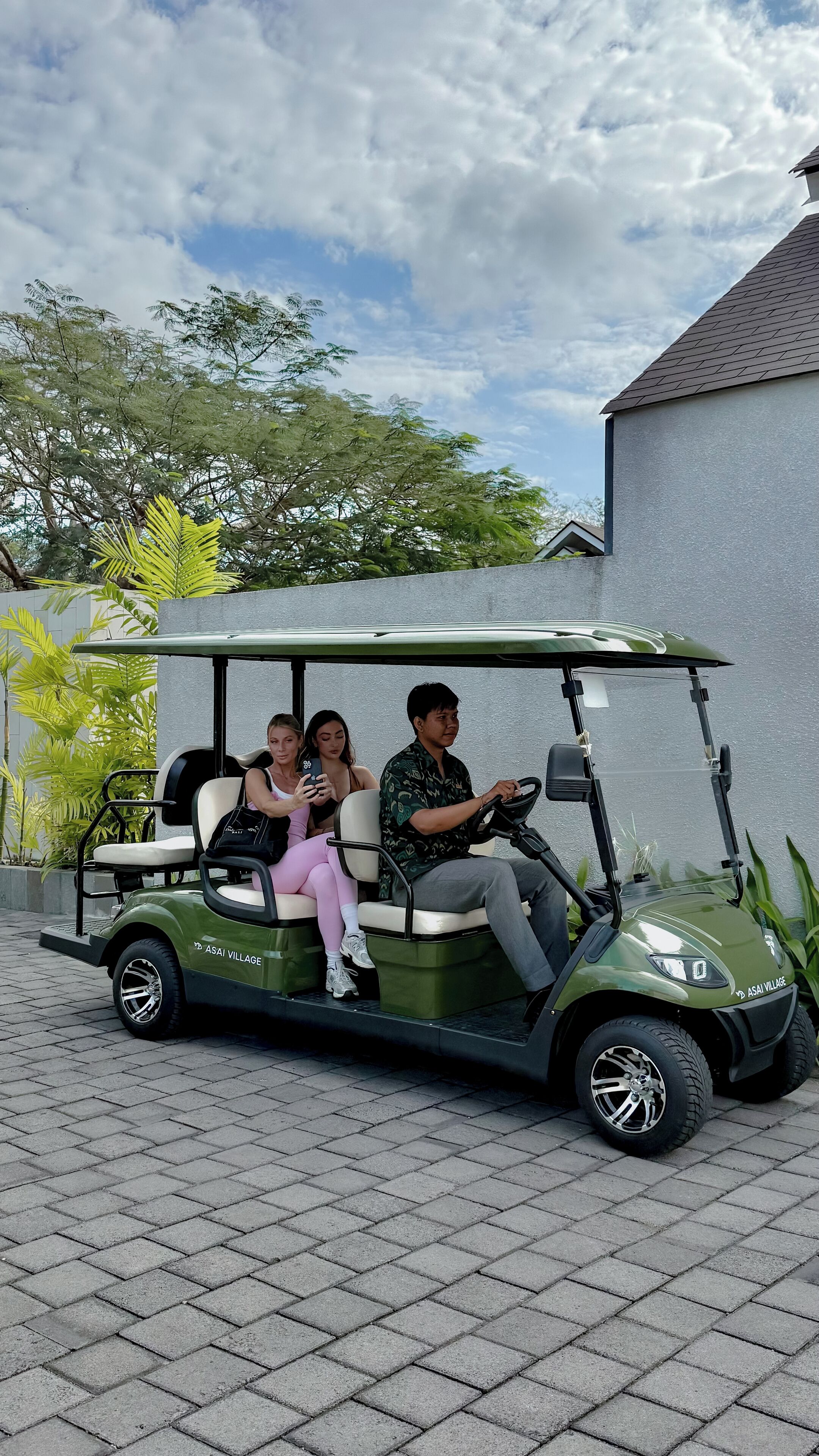 Une voiturette de golf avec trois passagers, garée sur une surface pavée, avec un bâtiment et des arbres en arrière-plan.