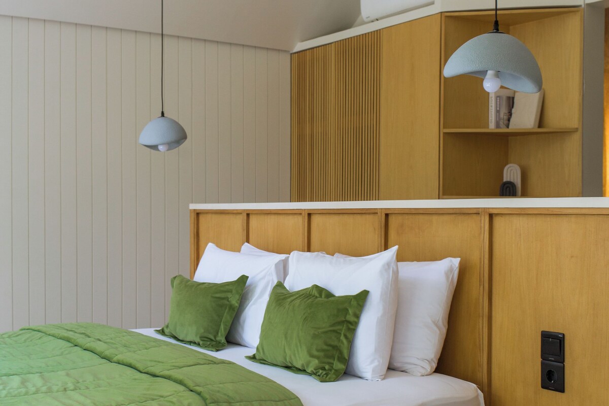 Un dormitorio moderno con cama, ropa de cama verde y blanca, un taburete pequeño y una lámpara colgante.