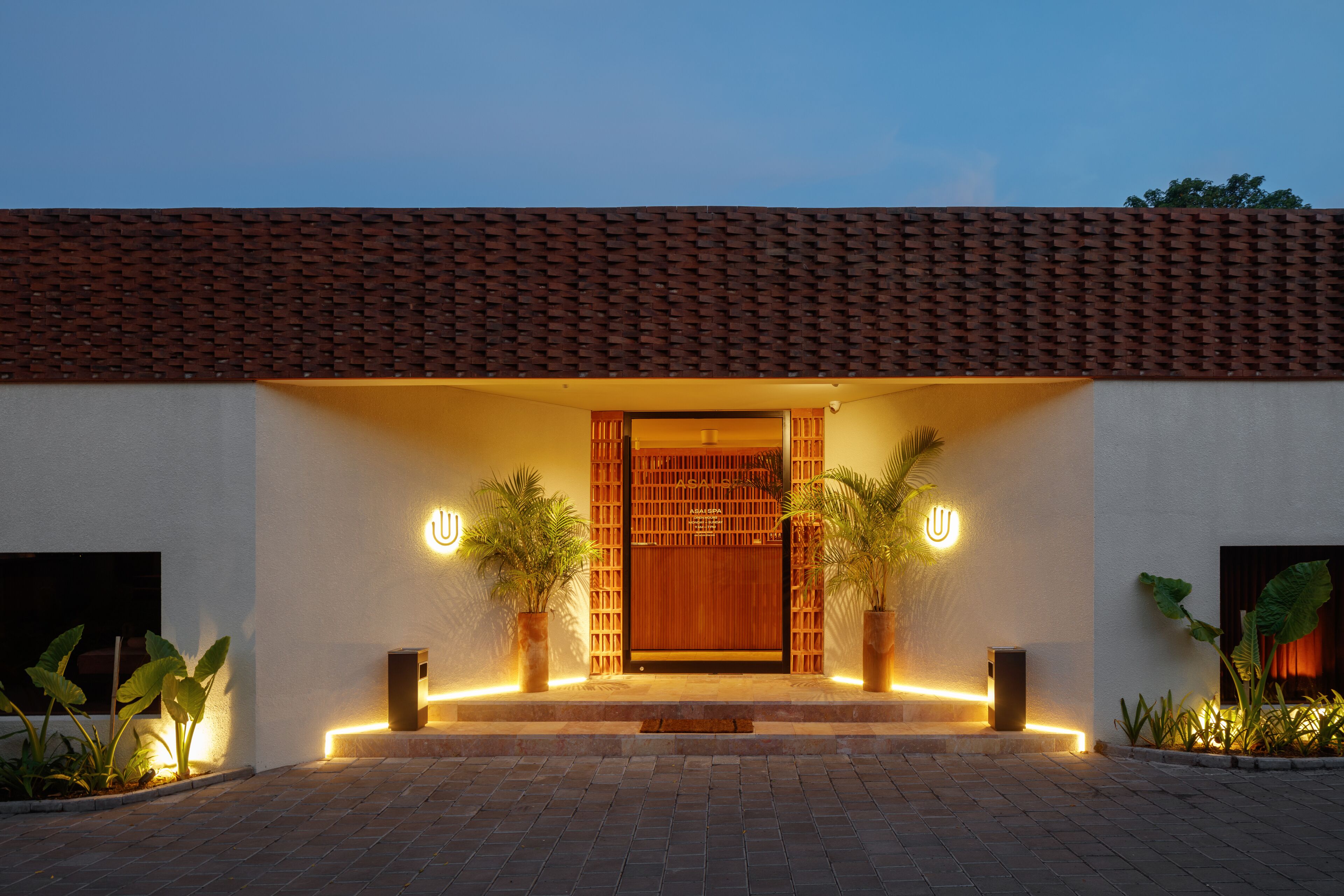 A modern building with a tiled roof, a wooden door, and two potted plants on either side.