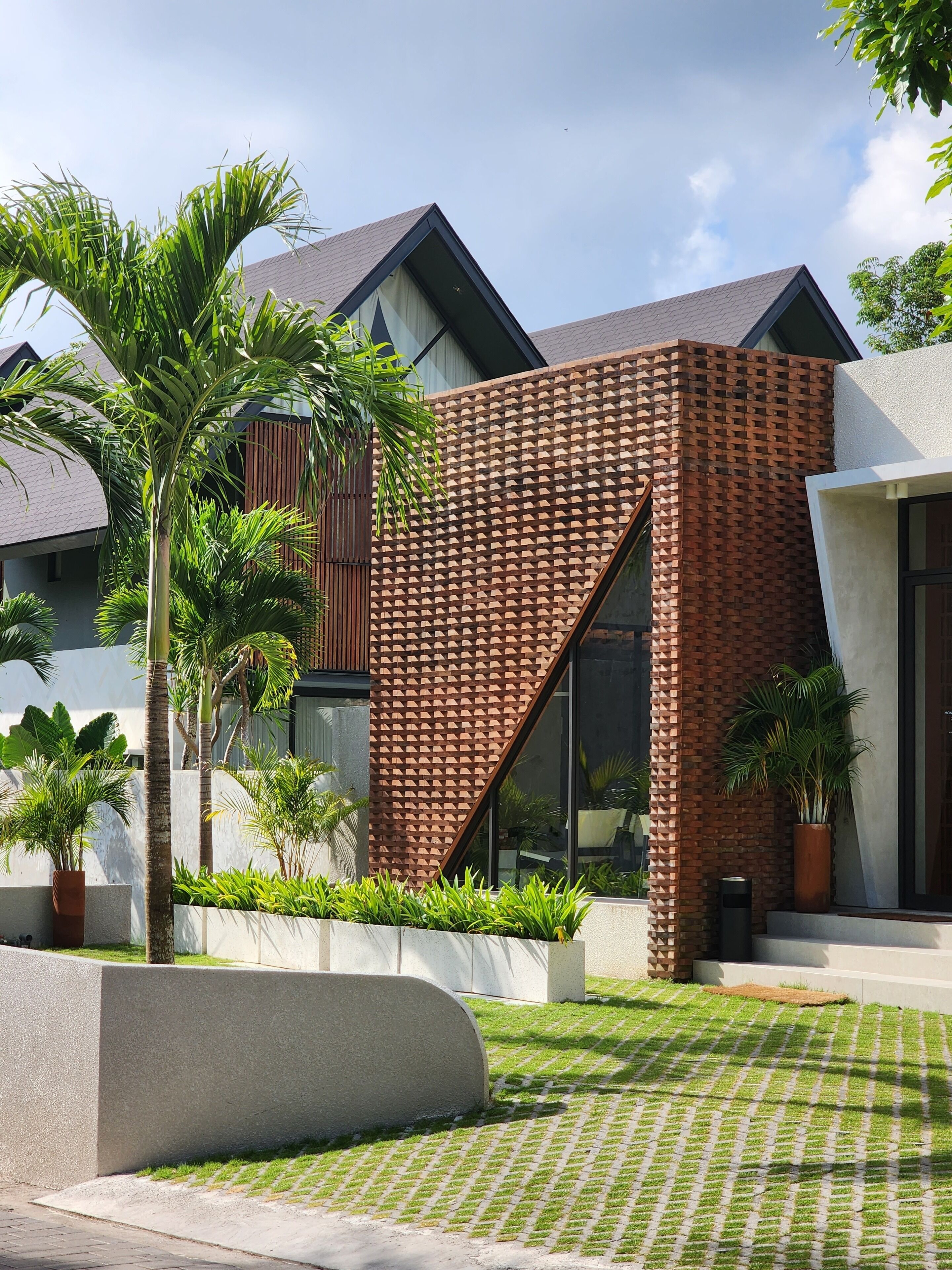 A modern building with a brick facade, large windows, and a well-maintained garden with palm trees.