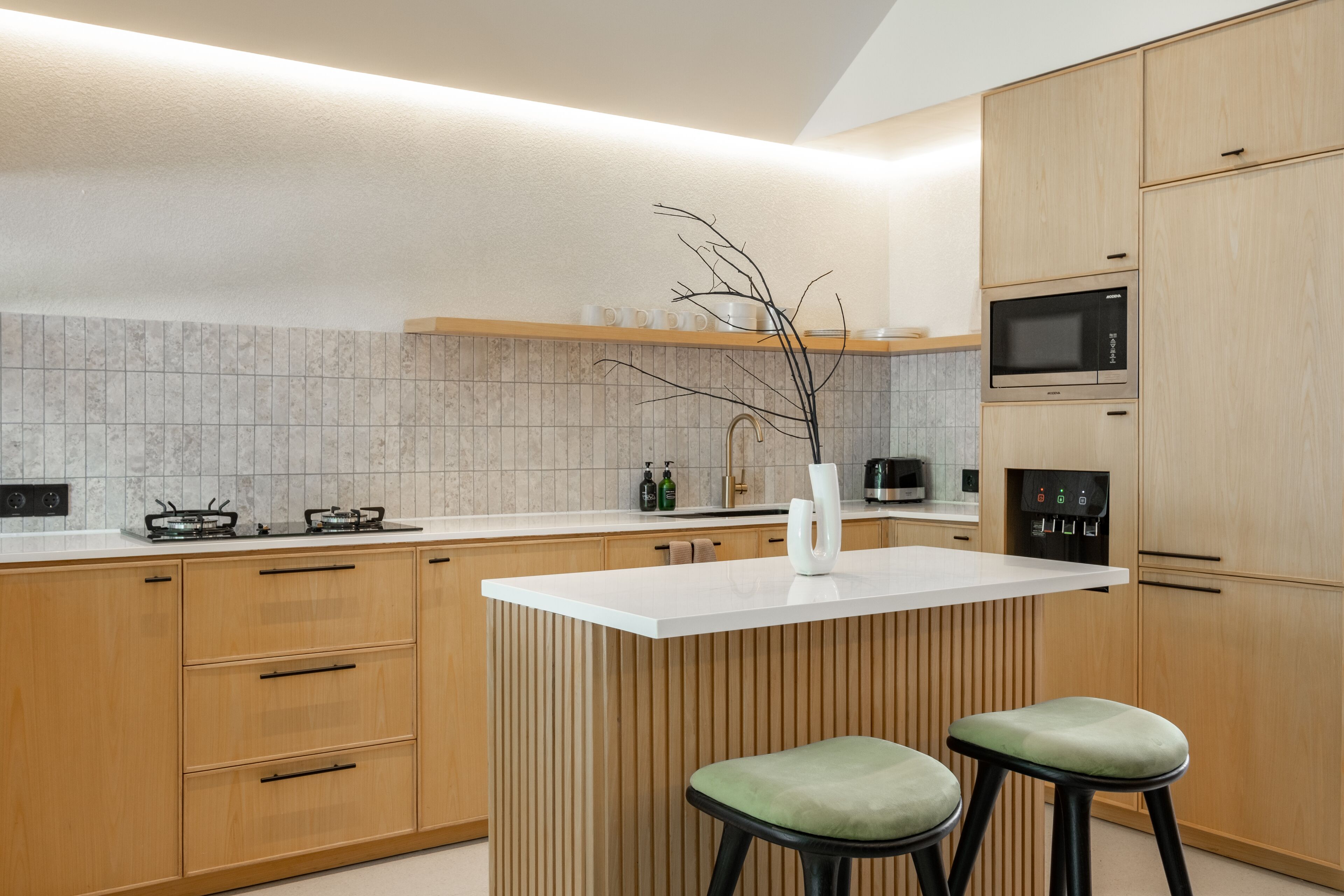 A modern kitchen with wooden cabinets, a white countertop, a built-in microwave, and two green bar stools.