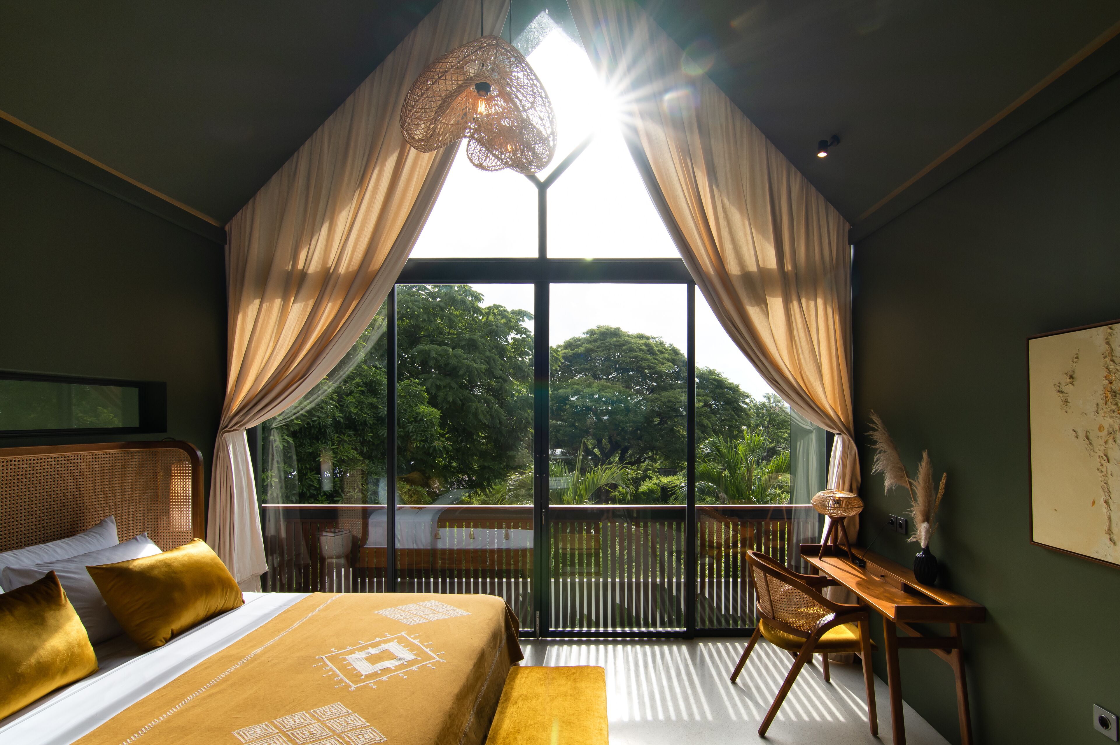 A bedroom with a large window, a bed with a patterned bedspread, a desk, and a chair.