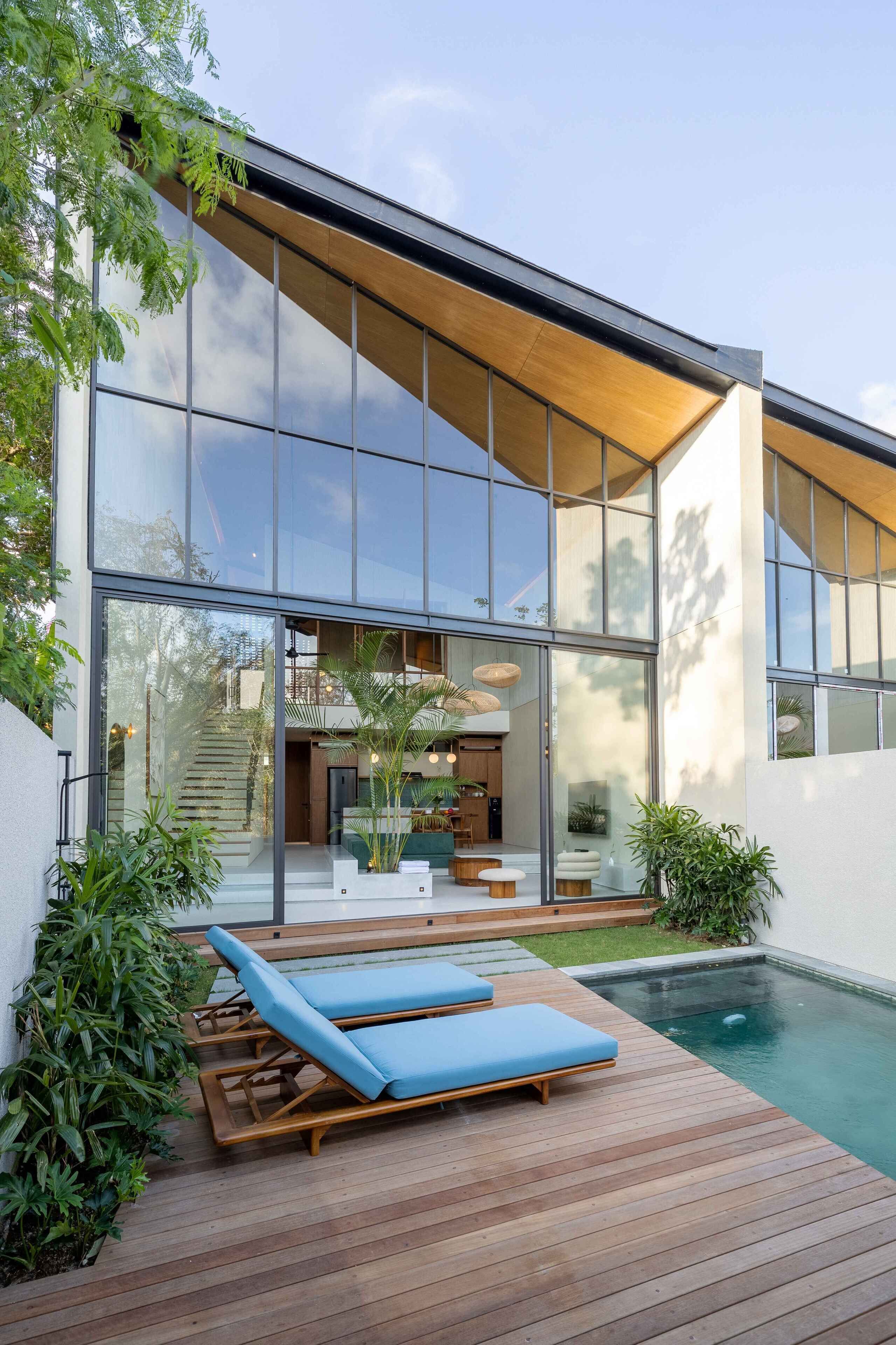 A modern house with a large glass facade, a wooden deck with lounge chairs, and a swimming pool.