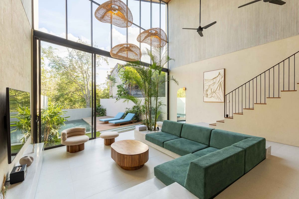 A modern living room with a green sofa, round wooden coffee table, and a TV on a stand. There are wicker lamps, a staircase, and a view of a pool and greenery outside.