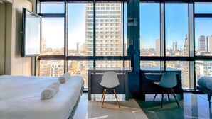 Een hotelkamer met een groot bed, een bureau en een stoel, met uitzicht op de stad door grote ramen.