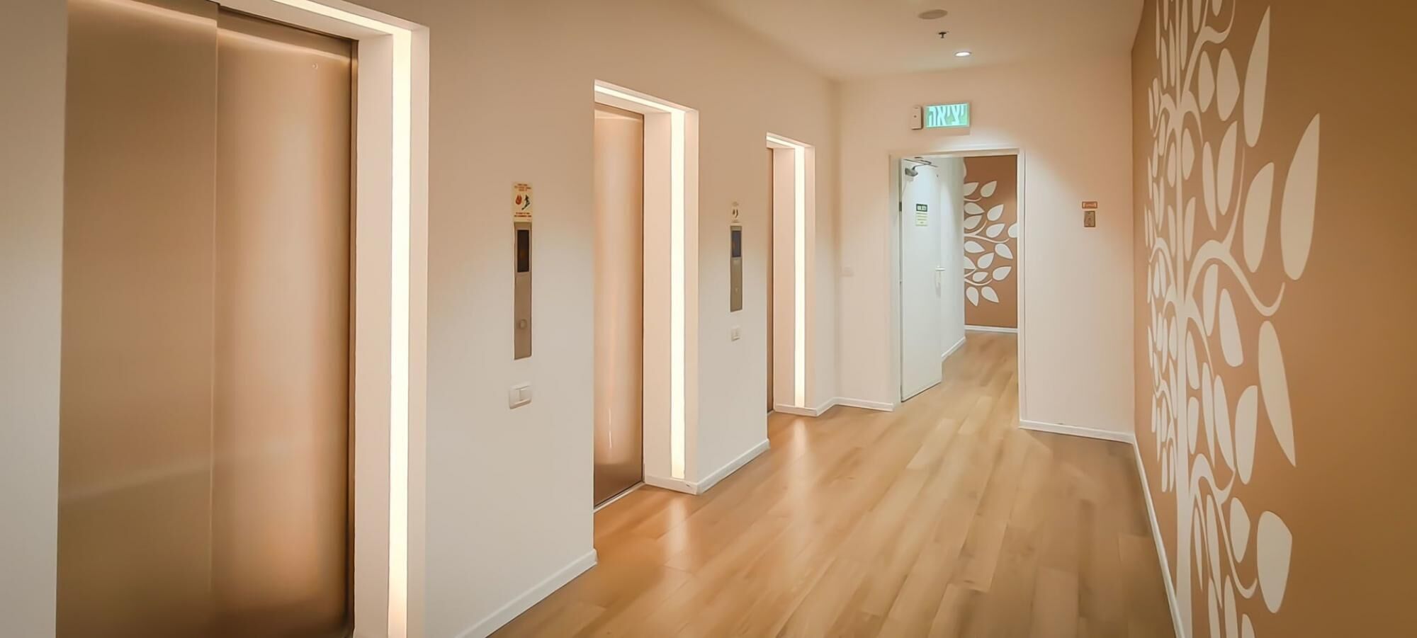 Ein Hotelgang mit Holzboden, weißen Wänden und einem dekorativen Wandmuster. Deutlich erkennbare Türen mit Notausgangsschildern und ein Aufzug sind vorhanden.