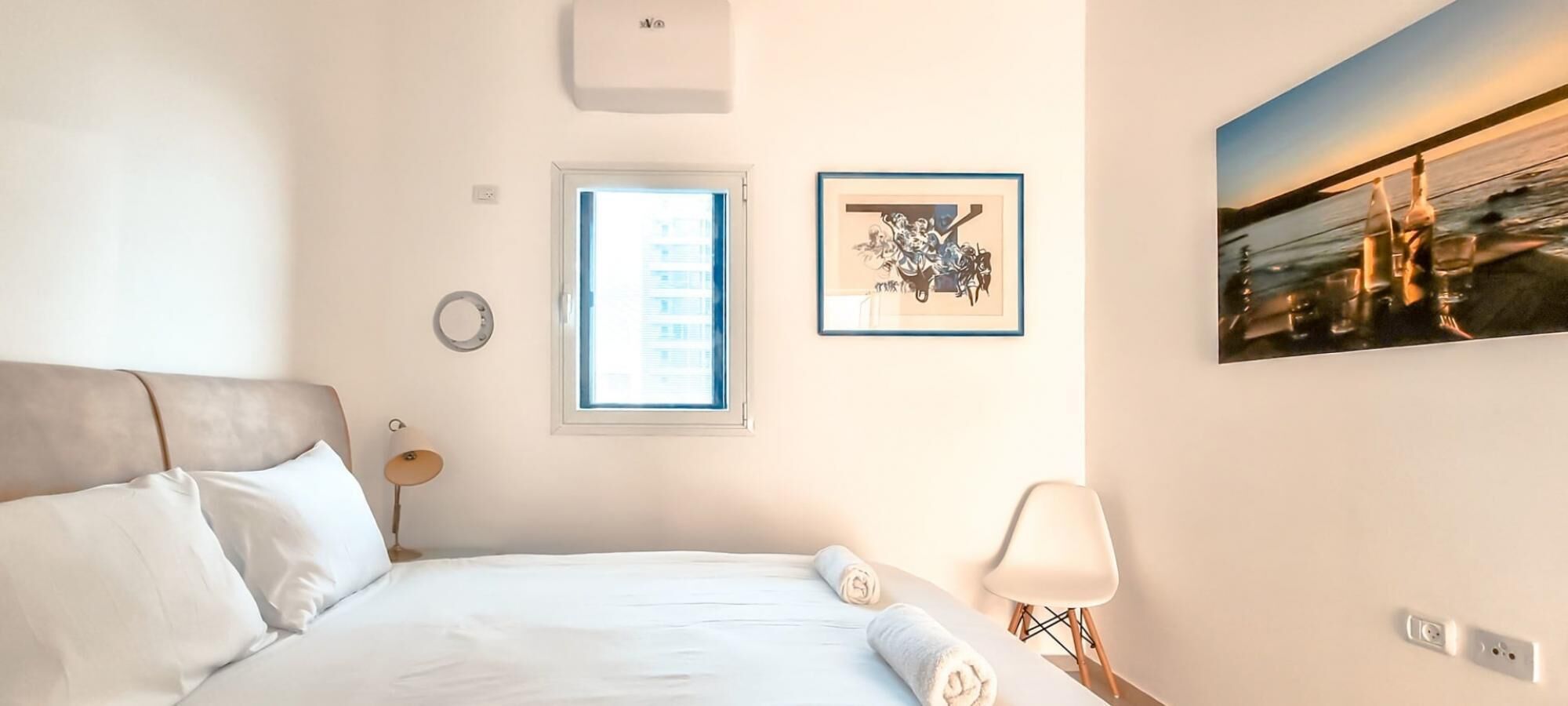 A hotel room with a bed, a window, a wall-mounted picture, and a nightstand with a lamp.