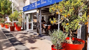 Un café avec une terrasse, des jardinières rouges et un auvent bleu orné d’une inscription.