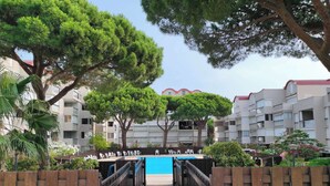 Un chemin en bois mène à une piscine entourée d’arbres, et à un immeuble à plusieurs étages doté de balcons.
