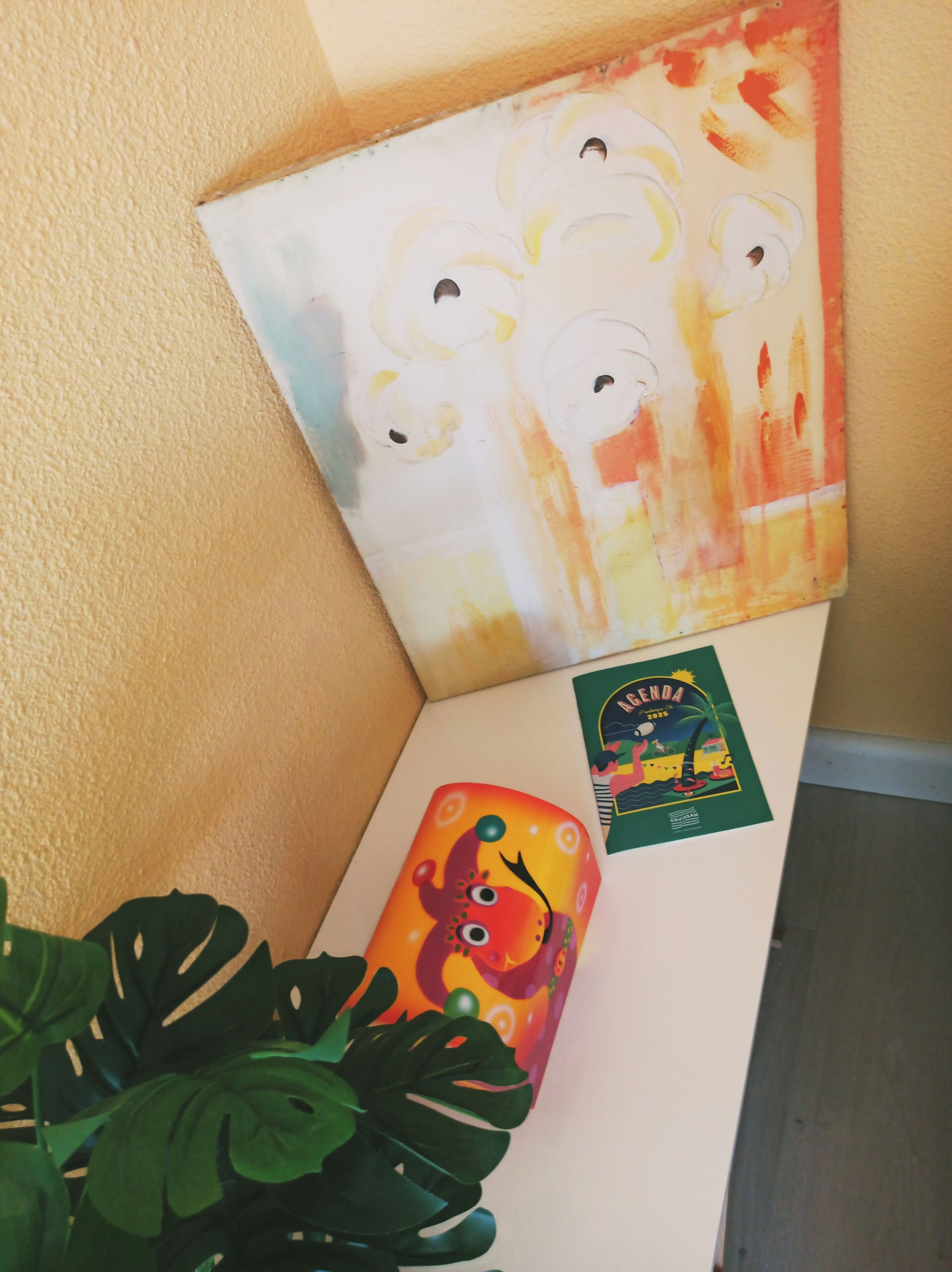 A corner of a room with a painting, a calendar, and a plant.