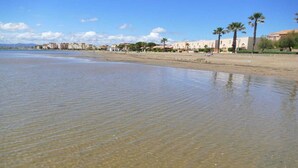 Une plage avec une eau claire et peu profonde, des palmiers et des bâtiments en arrière-plan.