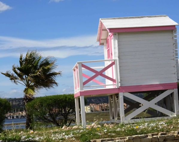 A beach hut with a white and pink exterior, situated on a grassy area with a palm tree and a body of water in the background.