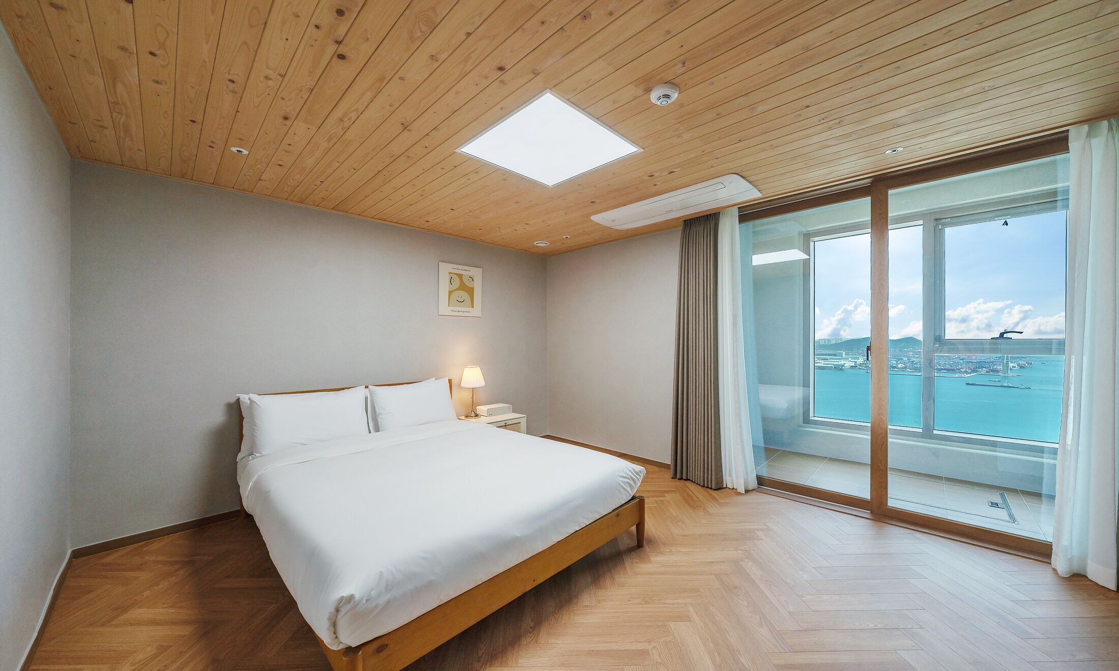 A bedroom with a bed, a nightstand, a lamp, a ceiling light, and a large window with a view of a body of water and a bridge.