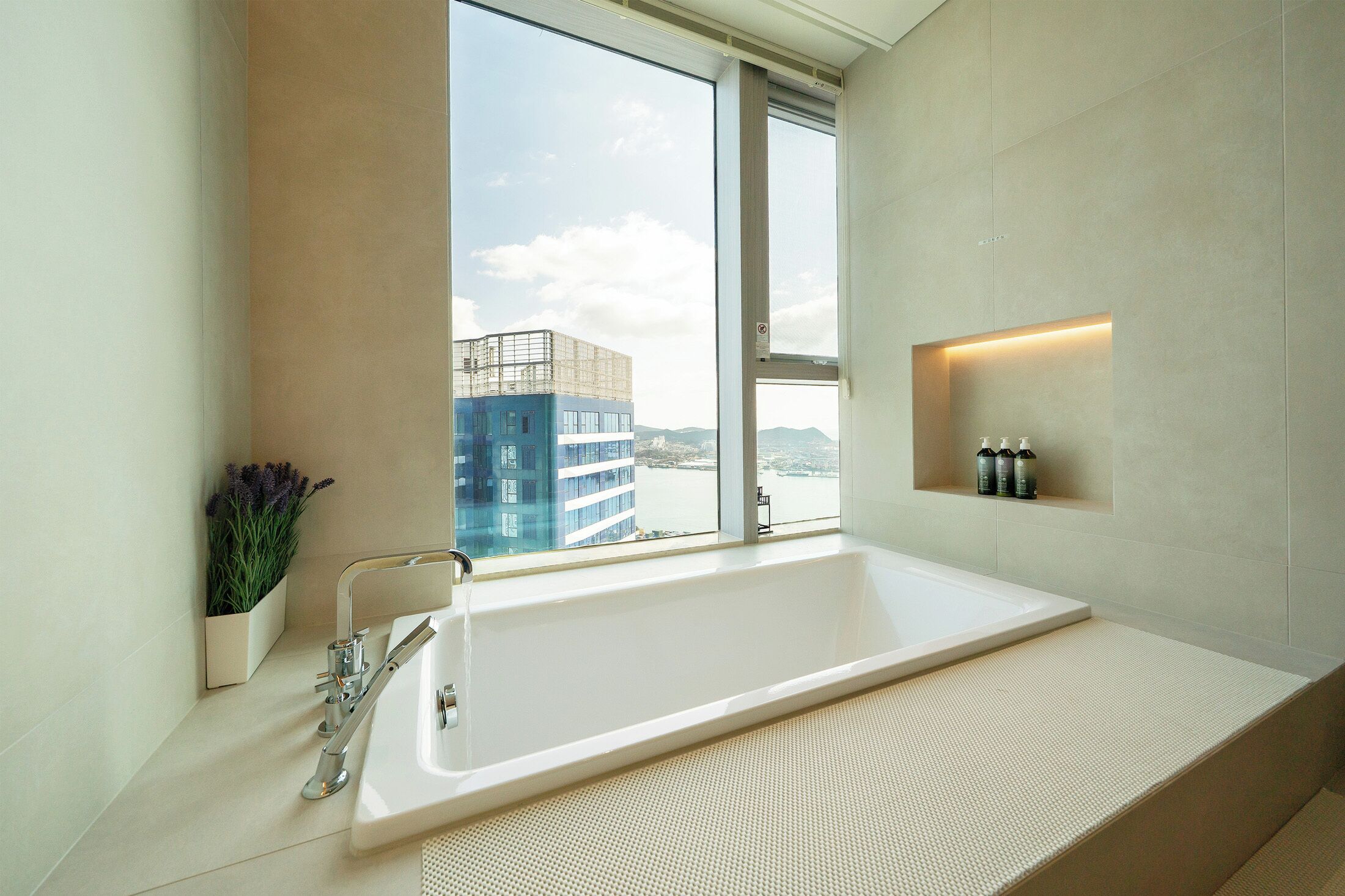 A modern bathroom with a large bathtub, a window with a view of buildings, and a shelf with toiletries.