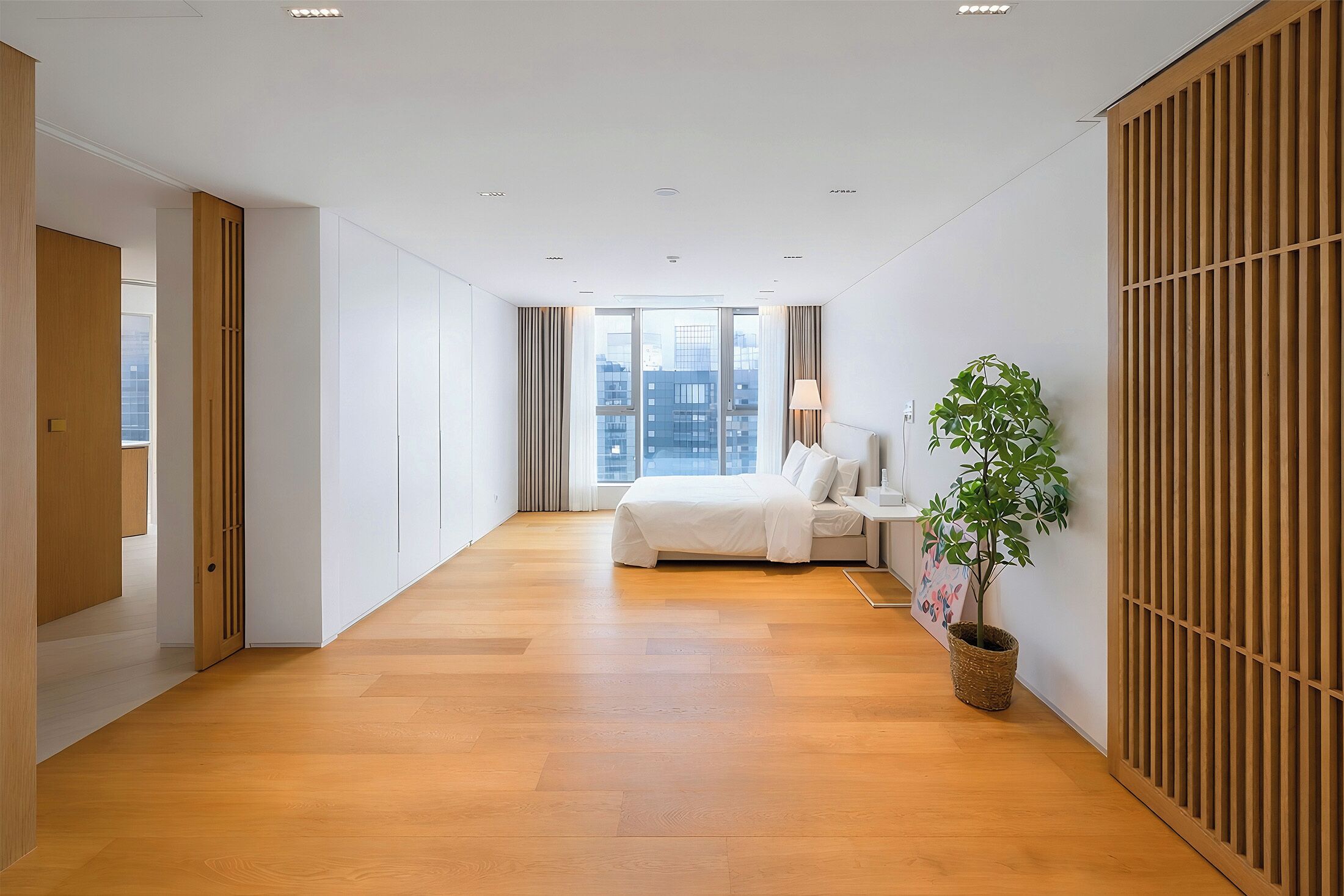 A modern hotel room with a wooden floor, a bed, a nightstand, a lamp, and a potted plant.