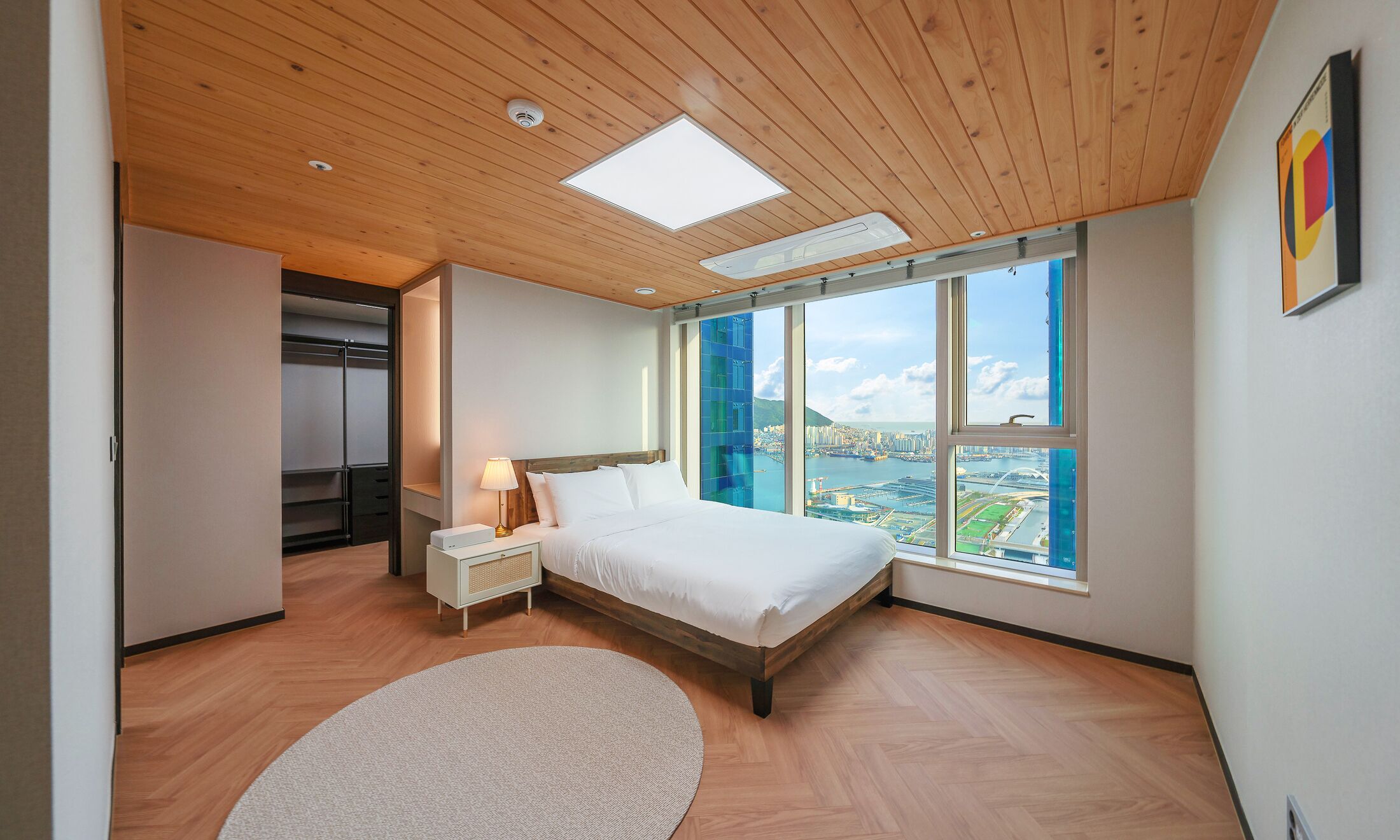 Un dormitorio moderno con una cama grande, una mesita de noche, una lámpara, un armario y un ventanal con vistas a la ciudad.