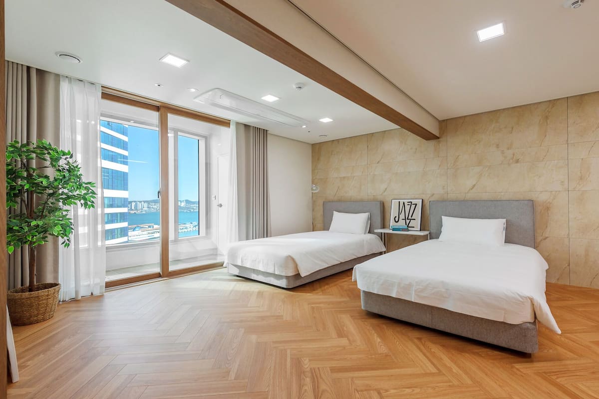 Quarto de hotel moderno com duas camas, uma janela grande com vista para a cidade, piso de madeira e uma planta em vaso.