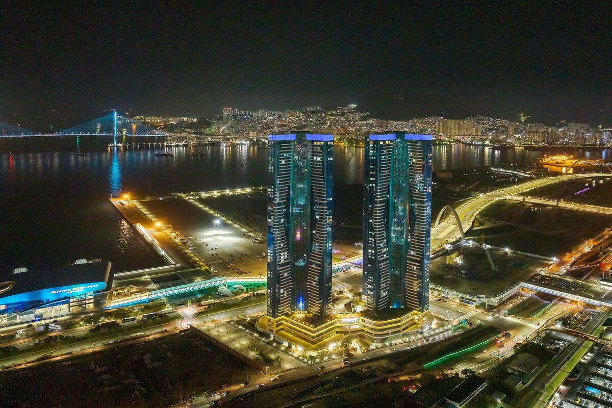 Vista aérea noturna de uma paisagem urbana com dois arranha-céus imponentes, uma ponte e um rio.