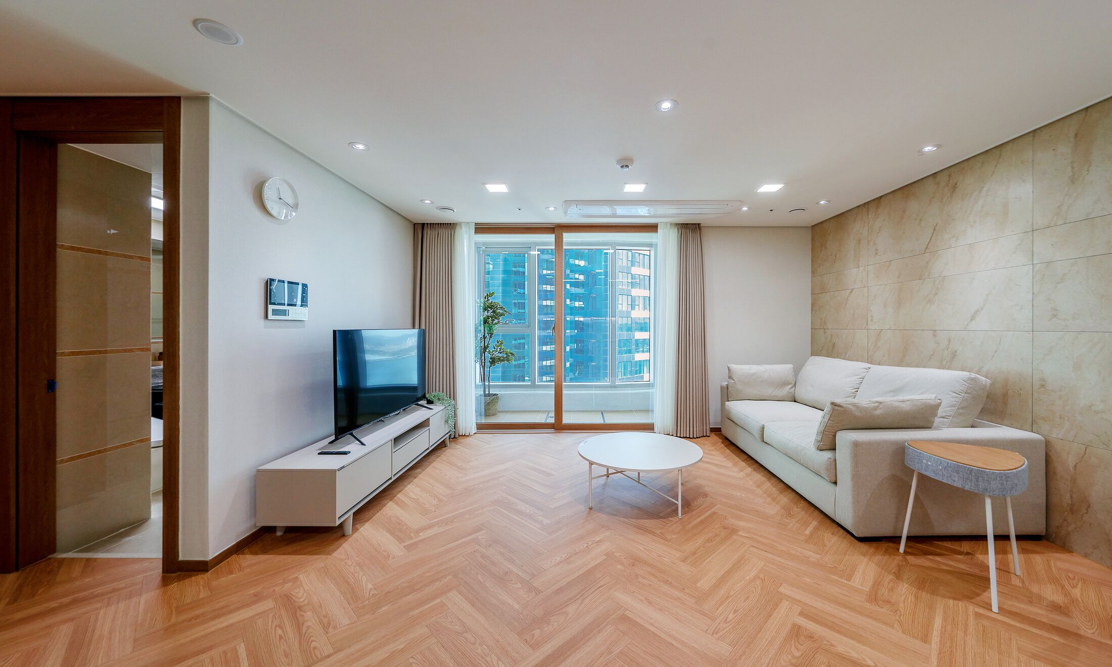 A modern living room with a flat-screen TV, a sofa, a coffee table, and a view of a cityscape through large windows.