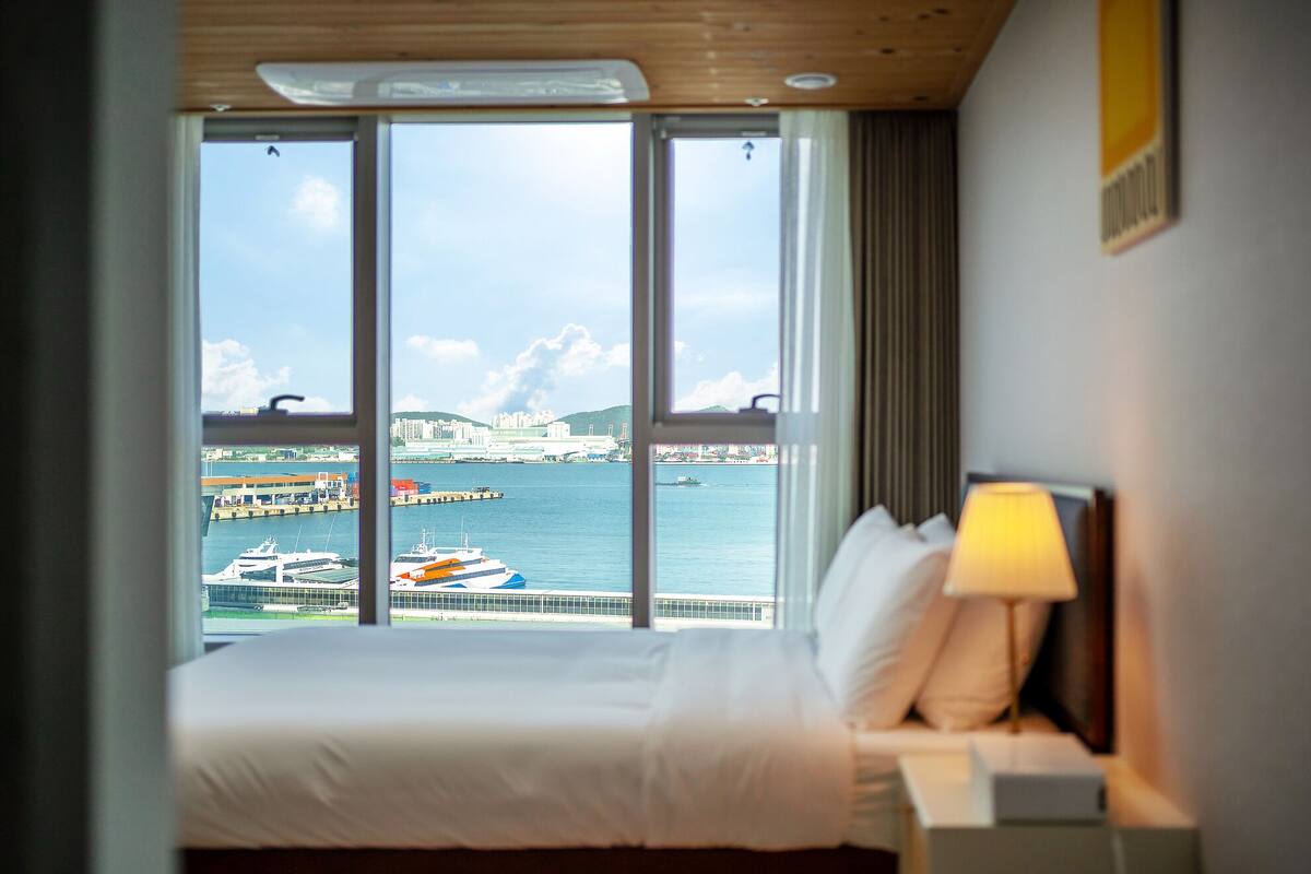 Quarto de hotel com uma grande janela que oferece vista para um porto com barcos e edifícios.