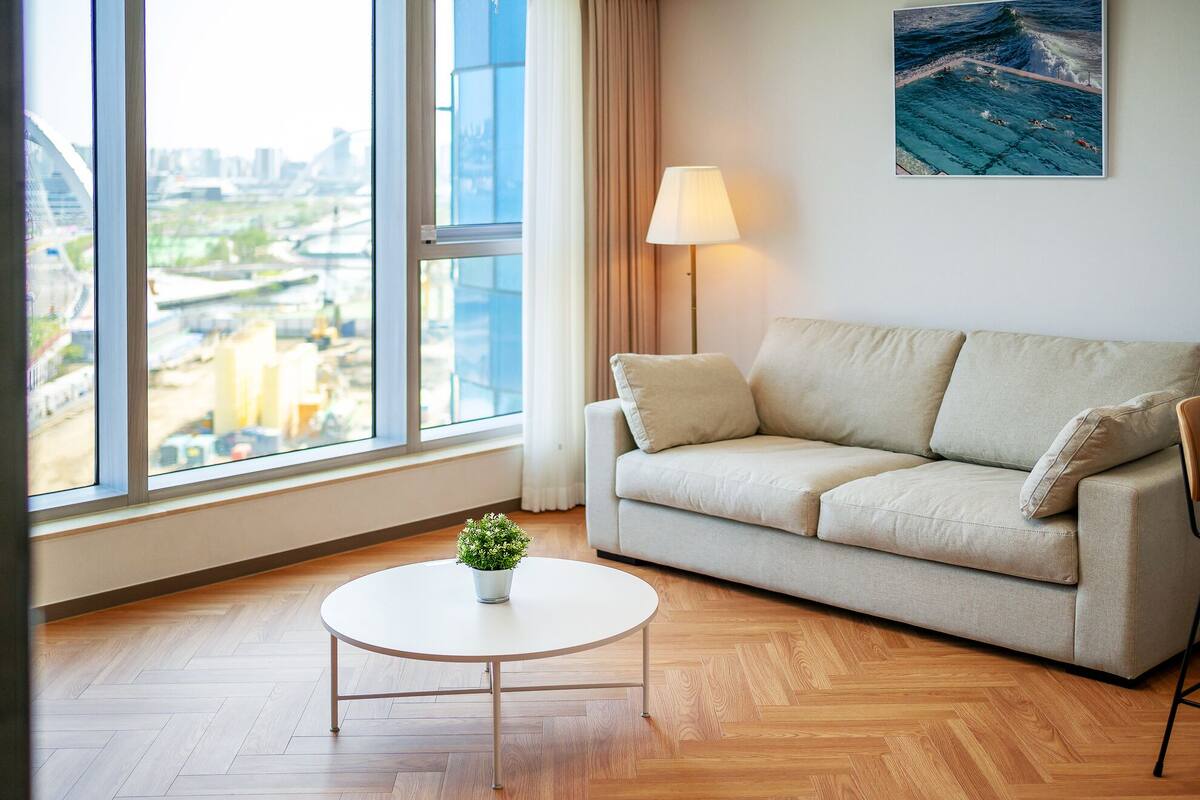 Uma sala de estar moderna com um sofá, uma mesinha redonda com uma planta e vista para a paisagem urbana através de grandes janelas.
