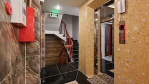 Un vestíbulo de hotel con una pared de mármol, un cuadro rojo de emergencia, una escalera y un ascensor.