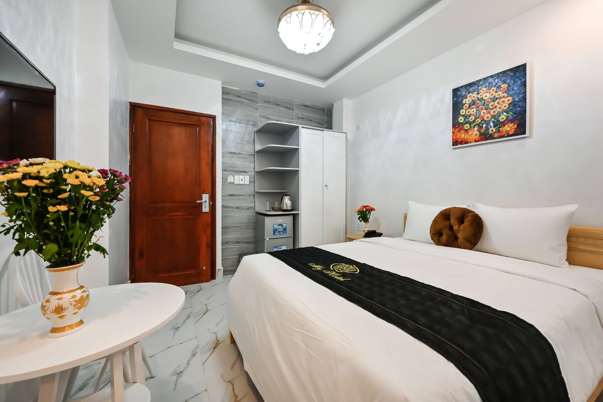 Habitación de hotel con una cama, una mesa redonda blanca con un florero, un armario, un cuadro en la pared y suelo de mármol.
