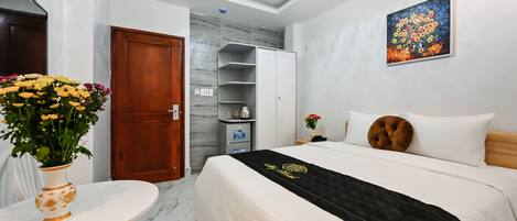 A hotel room with a bed, a white round table with a vase of flowers, a wardrobe, a painting on the wall, and a marble floor.