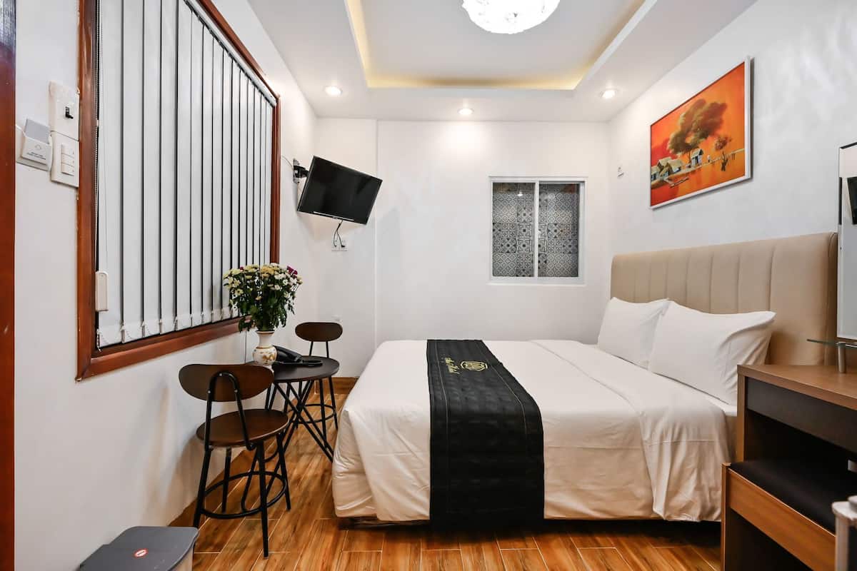 Habitación de hotel con una cama, un escritorio con dos sillas, una mesita con un jarrón de flores, una ventana con cortinas estampadas, un televisor montado y un cuadro en la pared.