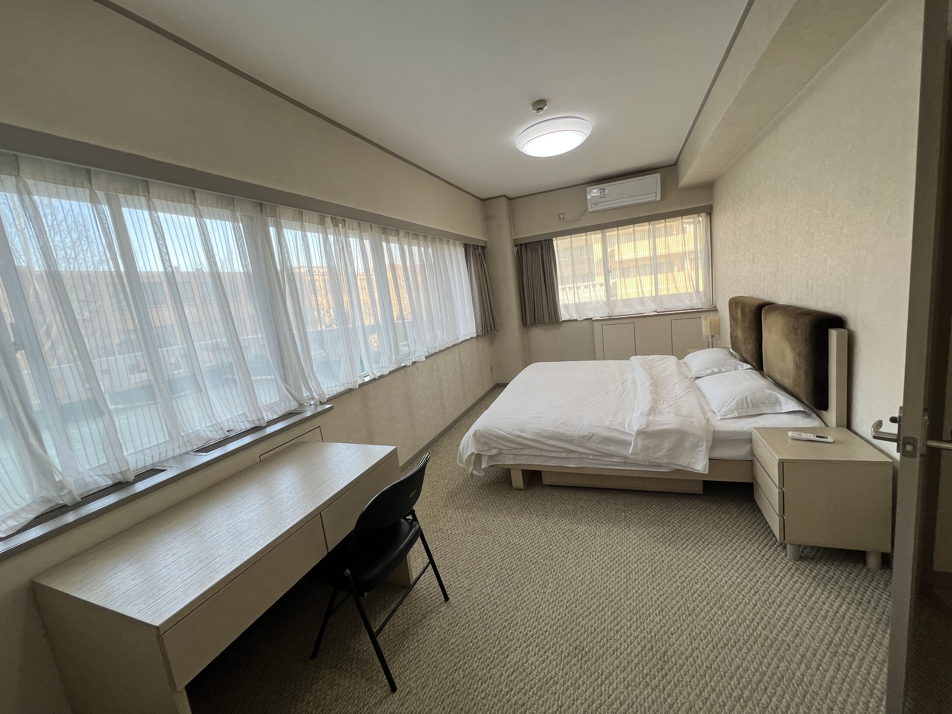 A hotel room with a bed, a desk, and a chair. There is a window with curtains, a bedside table, and a nightstand.