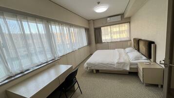 A hotel room with a bed, a desk, and a chair. There is a window with curtains, a bedside table, and a nightstand.