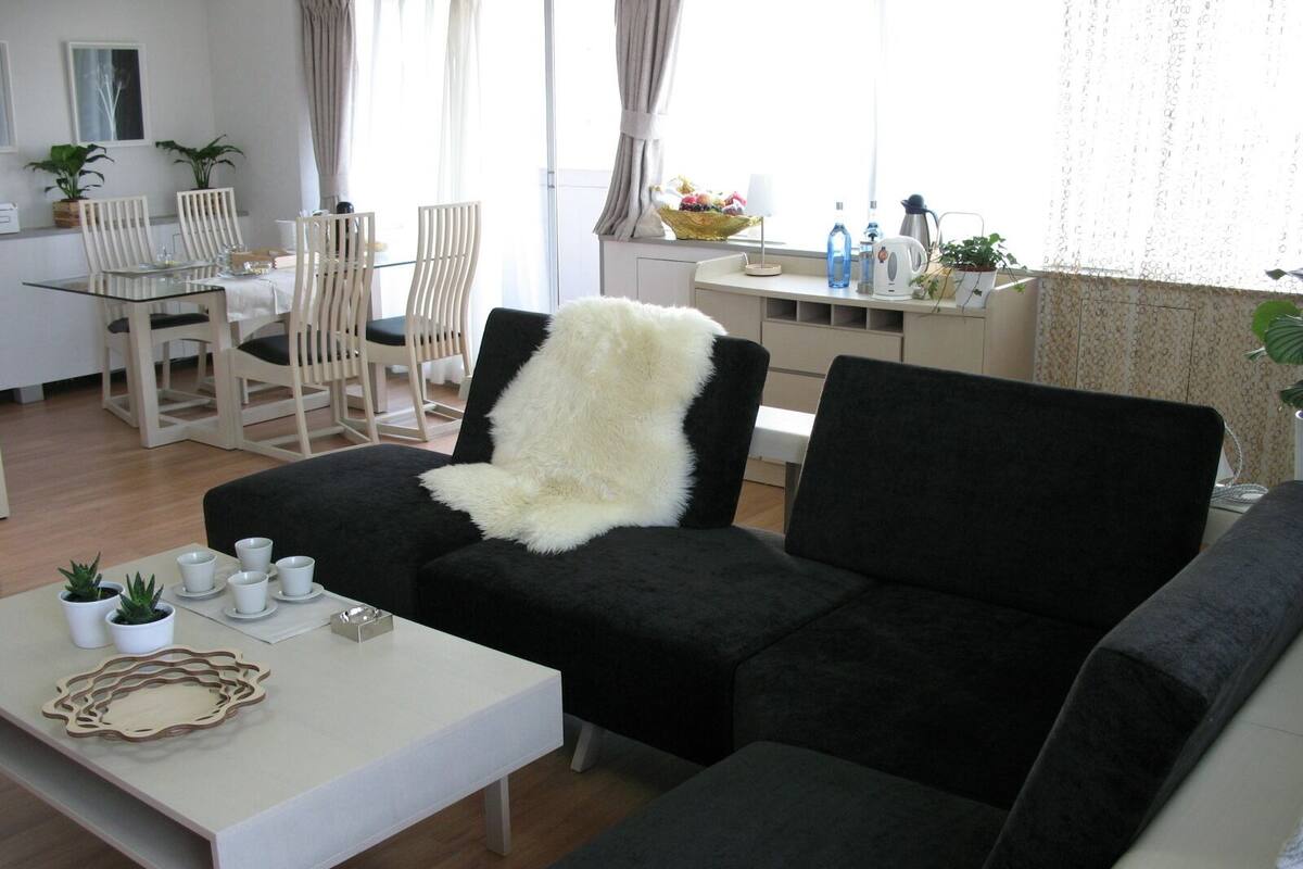 Ein modernes Wohnzimmer mit einer schwarzen Couch, einem weißen Couchtisch und einem Essbereich mit einem Glastisch und Holzsesseln.