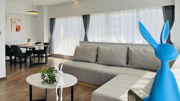 A modern living room with a grey sofa, a white table with a plant, and a blue rabbit decoration. There is a dining area with a table and chairs in the background. - Beijing Jiuxian Apartment (Beijing)